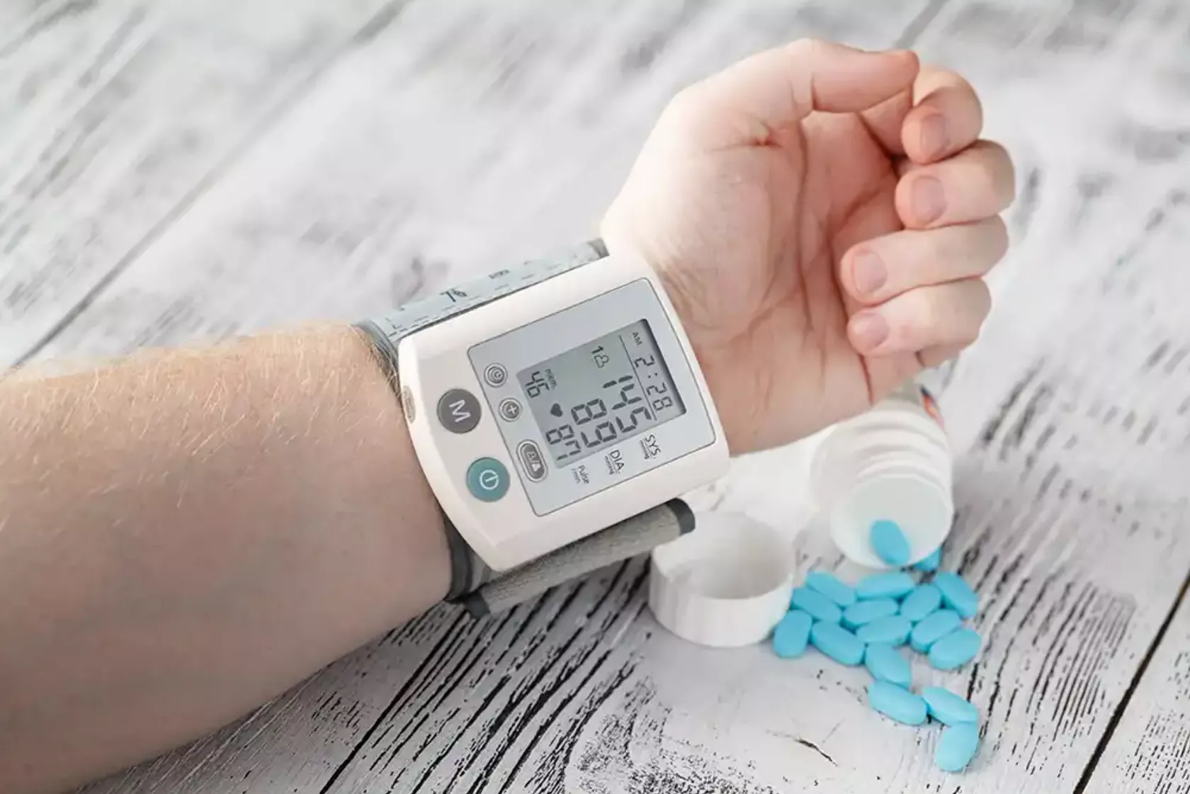Blutdruckmessgerät am Handgelenk eines Patienten und eines umgeschüttete Medikamentenpackung, blaue Pillen auf dem Tisch.