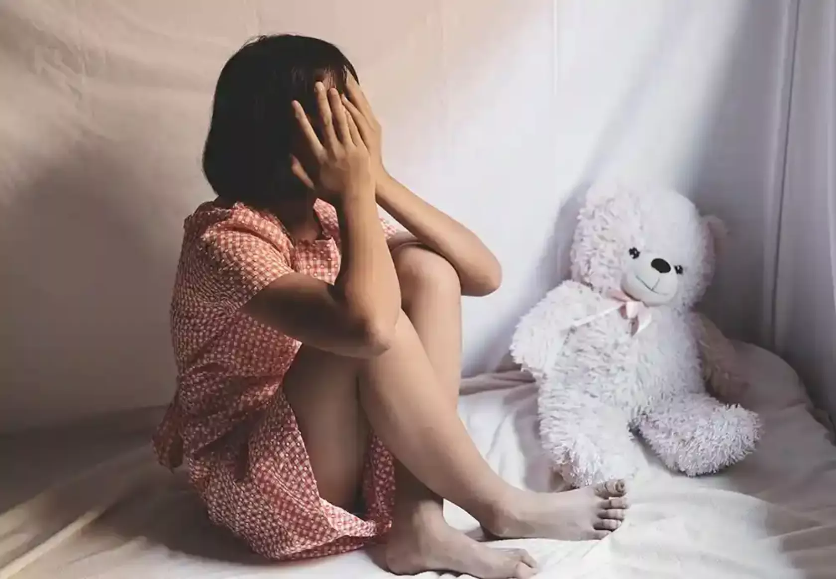 Mädchen sitzt mit verzweifeltem Ausdruck und den Händen vor dem Gesicht mit einem Teddybär im Bett.