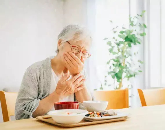 Seniorin sitzt vor ihrem Essen und hält die Hand vor den Mund.