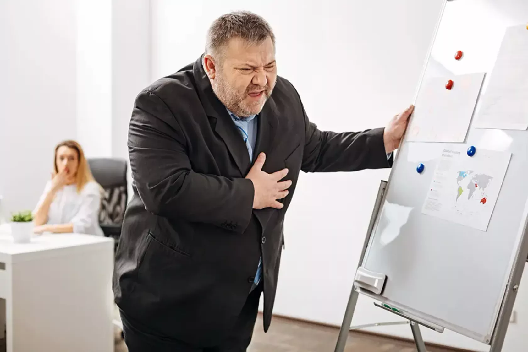 Mann greift sich während einer Präsentation im Büro mit schmerzerfülltem Gesichtsausdruck an die Brust.