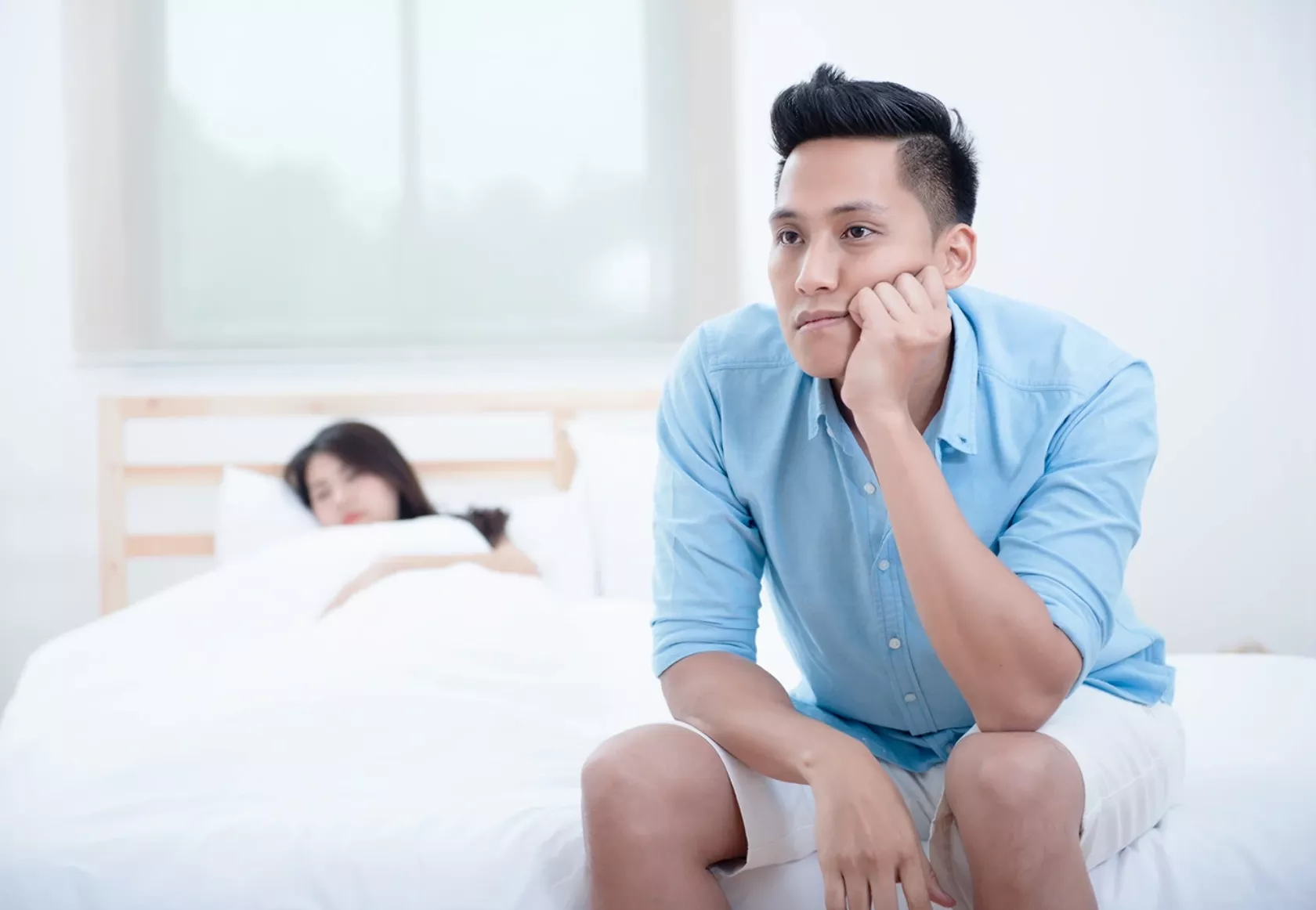 Junger Mann sitzt traurig am Rand des Bettes, in dem seine Partnerin liegt.