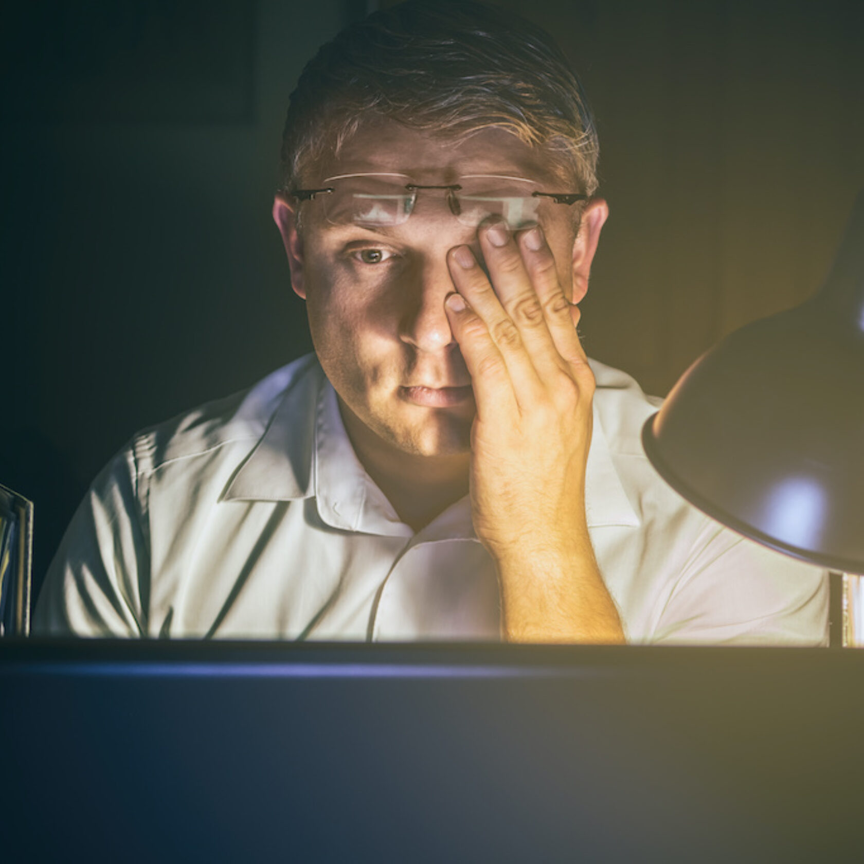 Mann im Dunklen bei Licht einer Schreibtischlampe wischt sich über das Auge