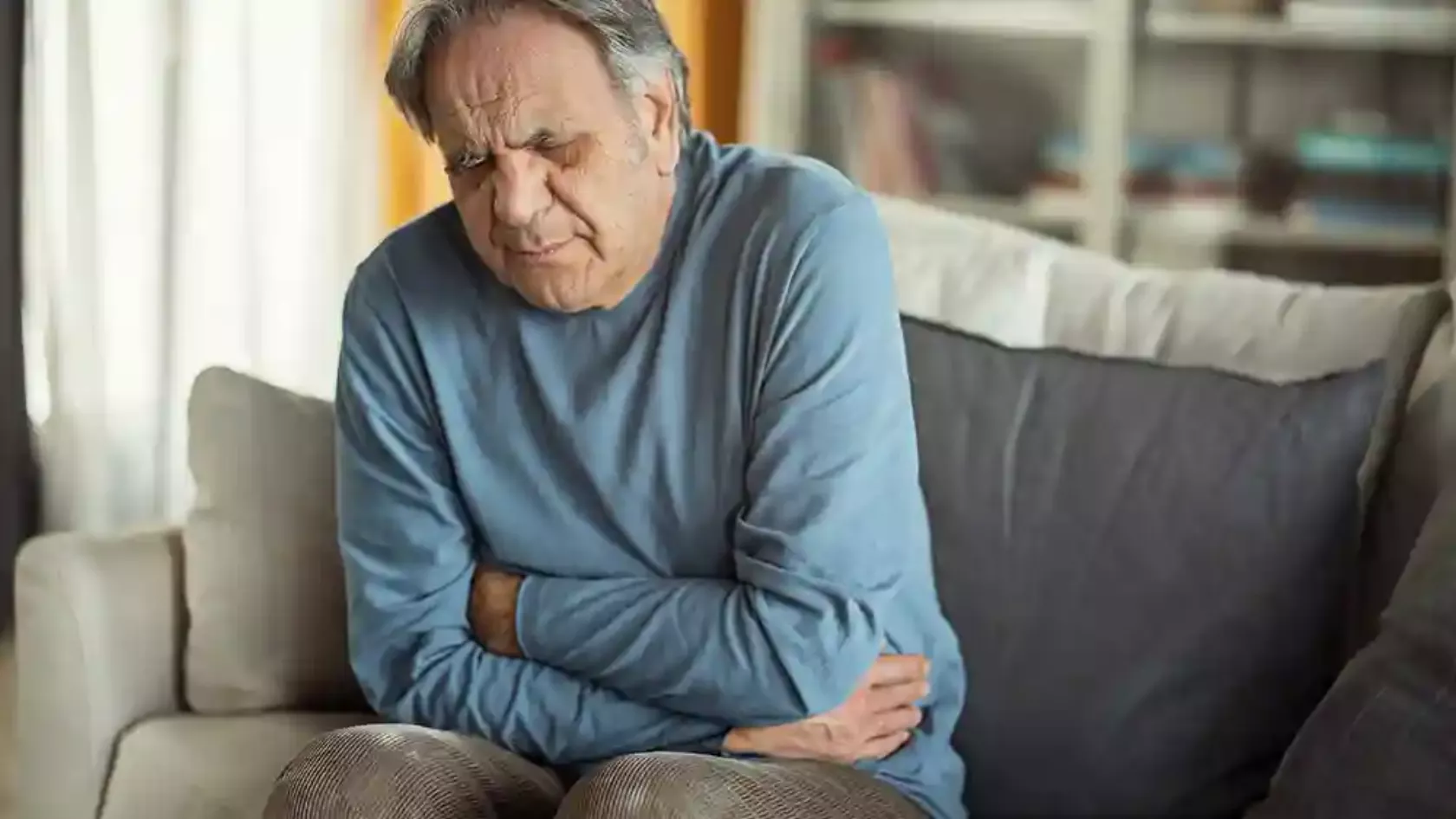 Älterer Mann sitzt auf dem Sofa und hält sich den Bauch, Gesicht mit schmerzhaftem Ausdruck.