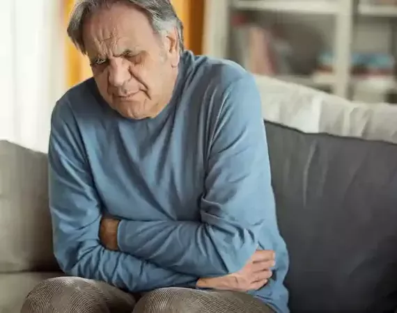 Älterer Mann sitzt auf dem Sofa und hält sich den Bauch, Gesicht mit schmerzhaftem Ausdruck.